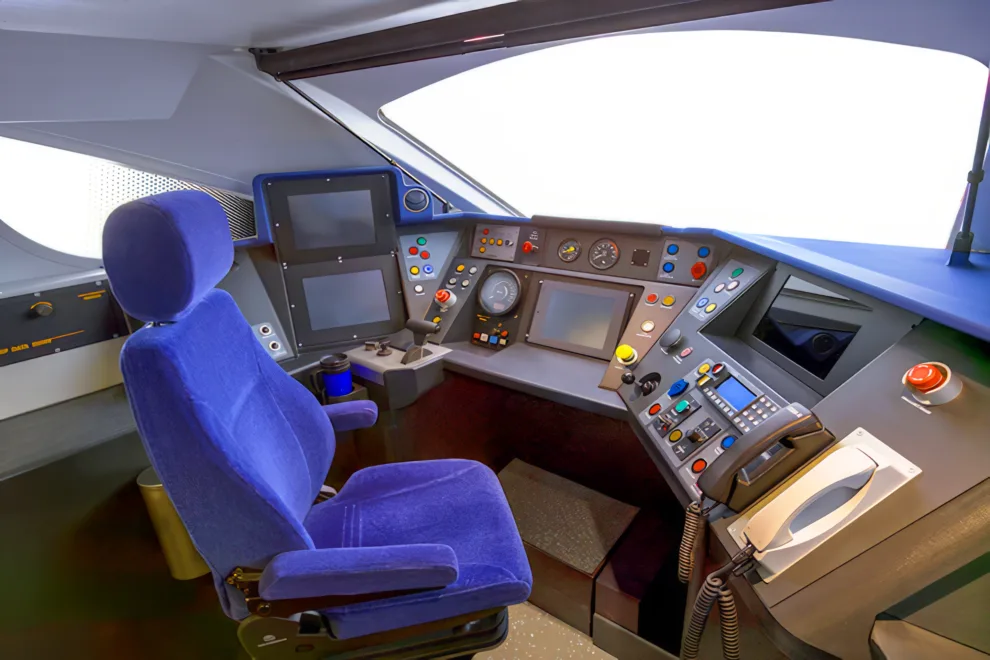 Photograph of the interior of a train cab, including a large blue chair in the foreground, in front of a series of dashboards with a variety of buttons, controls, screens, and instruments.