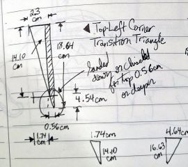 Photograph of a hand-drawn, dimensioned 2D drawing in a notebook, containing several triangles annotated with measurements.