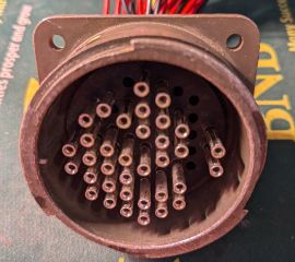 Inside view of a large circular connector, showing an irregular pattern of female pins, on top of a mousepad, with red, black, and yellow wires protruding from the back