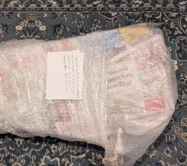 Bubble wrap and newspaper-wrapped parcel on a rug, including a hand-written label describing the packed PBC part within.